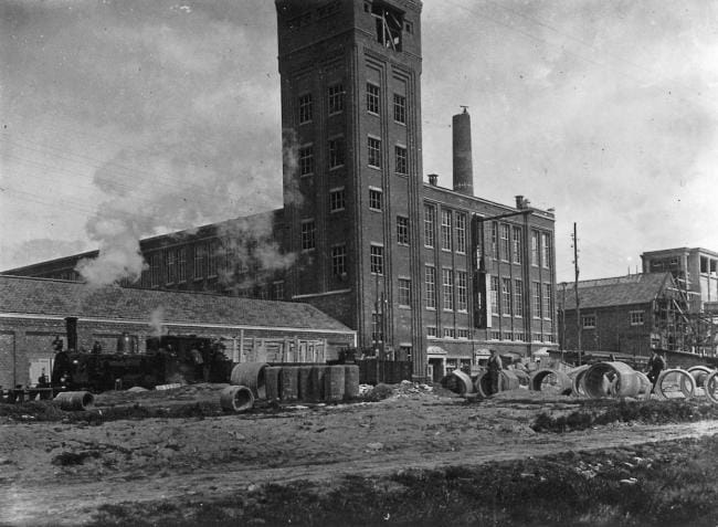 De textielfabriek van Van Heek in Losser in de jaren twintig. De fabriek is nog in aanbouw en krijgt bouwmateriaal per spoor aangevoerd. Dat gebeurt met een stoomlocomotief van de serie 7700. De fabriek bezat een eigen spooraansluiting en zou voor de reactivering van de tramlijn van grote historische betekenis blijken. Foto: Historische Kring Losser.